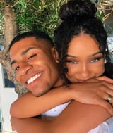 Duas pessoas posando juntas ao ar livre, uma abraçando a outra por trás perto de uma árvore