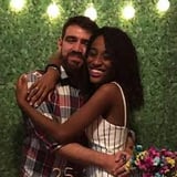 Duas pessoas sorrindo para a câmera, de braços dados em frente a parede de cerca verde e luzes decorativas